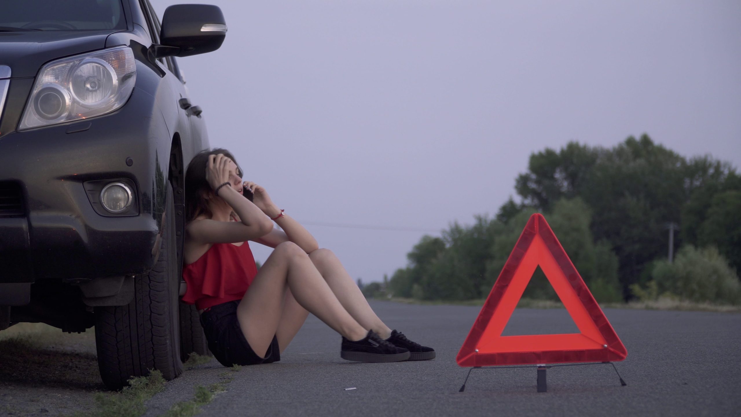 Woman sitting against her broken down car on the side of the road
